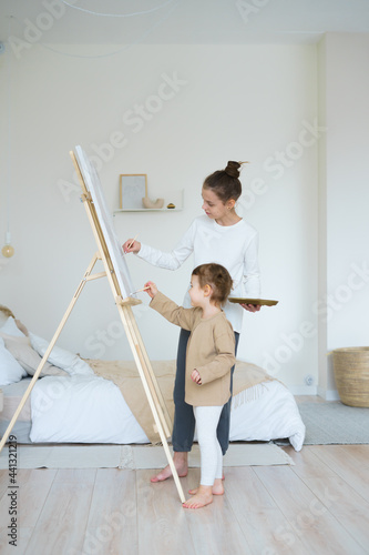 day apartment two girls sisters in home clothes are standing near an easel and drawing a picture