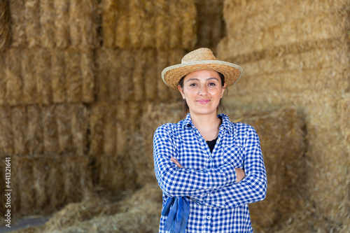 Portrait of successful young Hispanic female farmer posing with crossed arms near straw stack after work at farm, smiling confidently