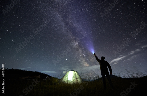 Back view of male traveler illuminating beautiful starry sky. Man hiker shining flashlight into night starry sky with Milky Way near illuminated camp tent. Concept of travelling and night camping.