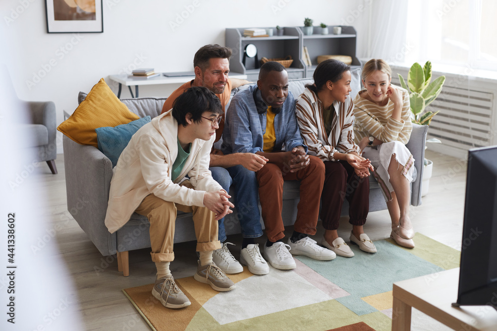 Fototapeta premium Full length view at multi-ethnic group of friends watching TV at home together while sitting in row on sofa, copy space