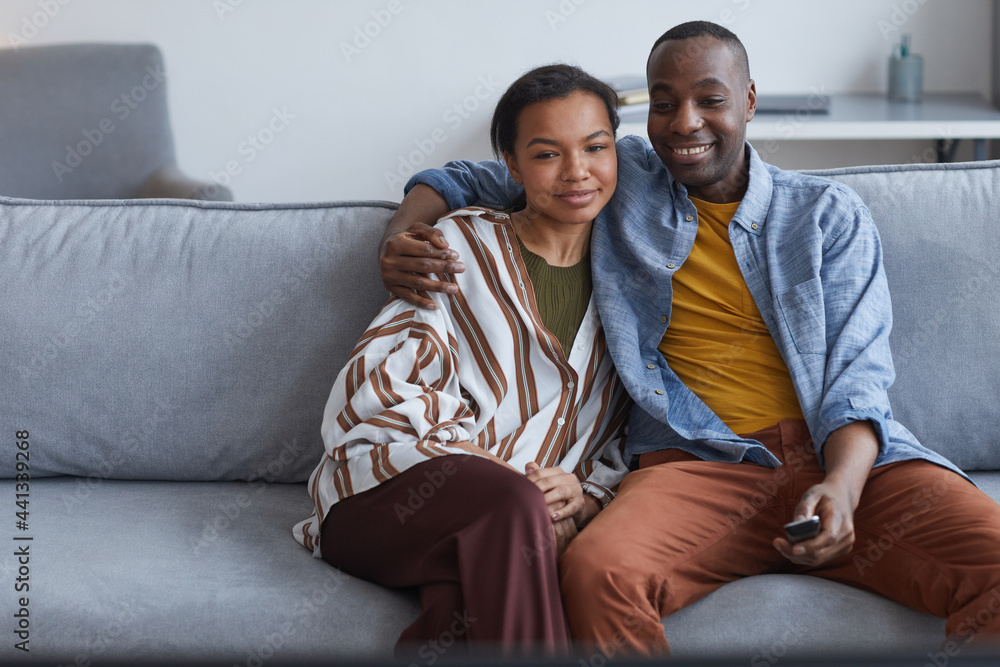 Portrait of young African-American couple watching TV or movies together while sitting on sofa at home, copy space