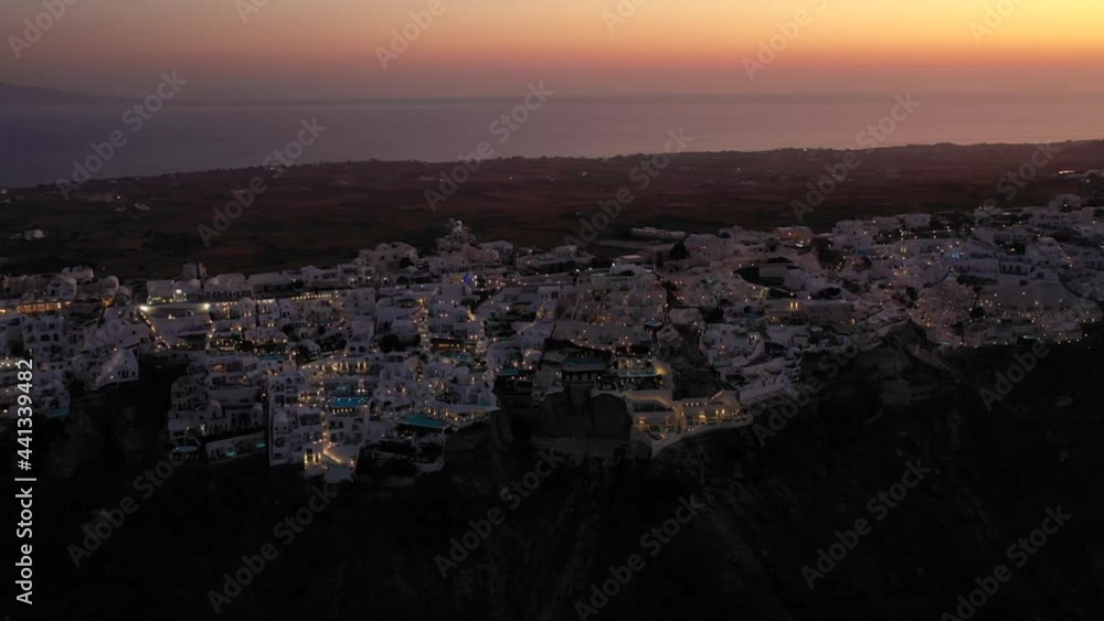Flight over of traditional terraced white villas in Oia town at sunset in the dark evening, Santorini, Greece. Aerial shot