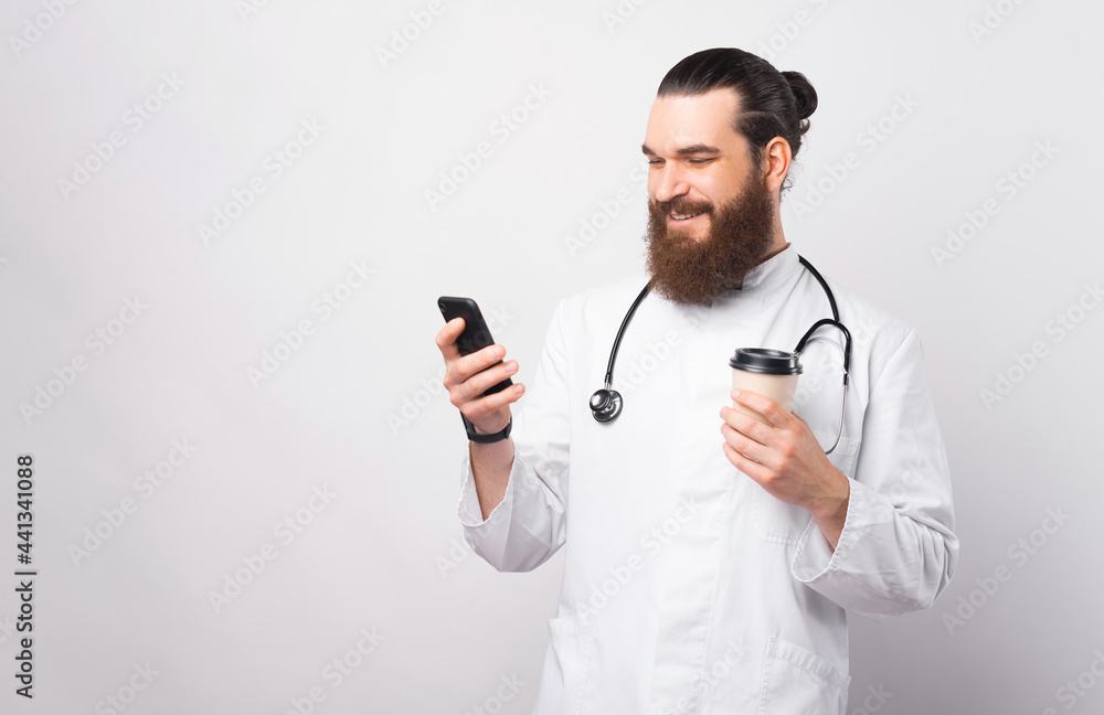 Photo of cheerful smiling young doctor man using smartphone and holding paper cup of coffee Photo of cheerful smiling young doctor man using smartphone and holding paper cup of coffee
