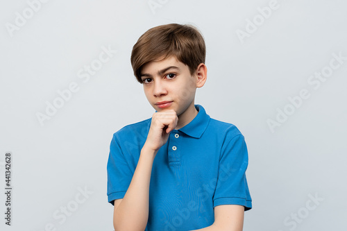 Portrait of a thoughtful boy holding hand on chin and looking at camera while standing isolated on light gray background © AstiMak