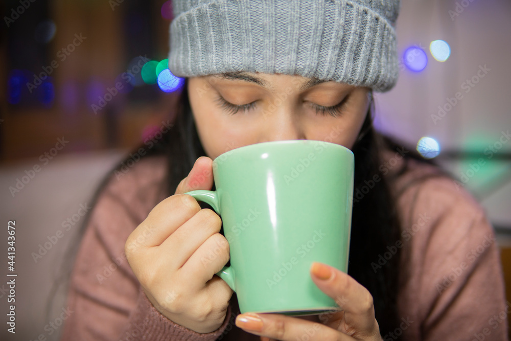 Indoor image of an Asian, Indian beautiful, serene young woman drinking coffee winter season at home. She is wearing warm clothes, sweater and a woolen cap. Indoor image of an Asian, Indian beautiful, serene young woman drinking coffee winter season at home. She is wearing warm clothes, sweater and a woolen cap.