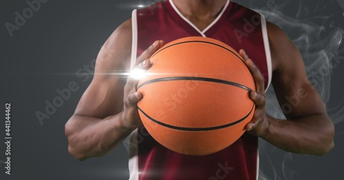 Mid section of male basketball player holding ball against smoke and light spot on grey background