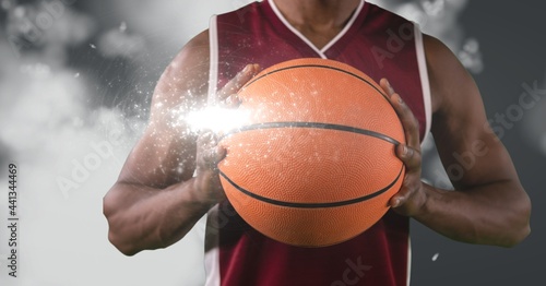Mid section of male basketball player holding basketball against light spot on grey background