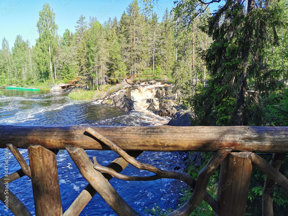 View from the observation deck of the picturesque waterfall on the Tokhmayoki river in Karelia surrounded by trees on a clear summer morning. View from the observation deck of the picturesque waterfall on the Tokhmayoki river in Karelia surrounded by trees on a clear summer morning.
