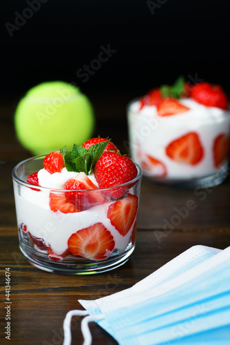 Whipped cream and strawberries served in a glass. Dark wooden table, tennis ball in bokeh, surgical masks, high resolution photo