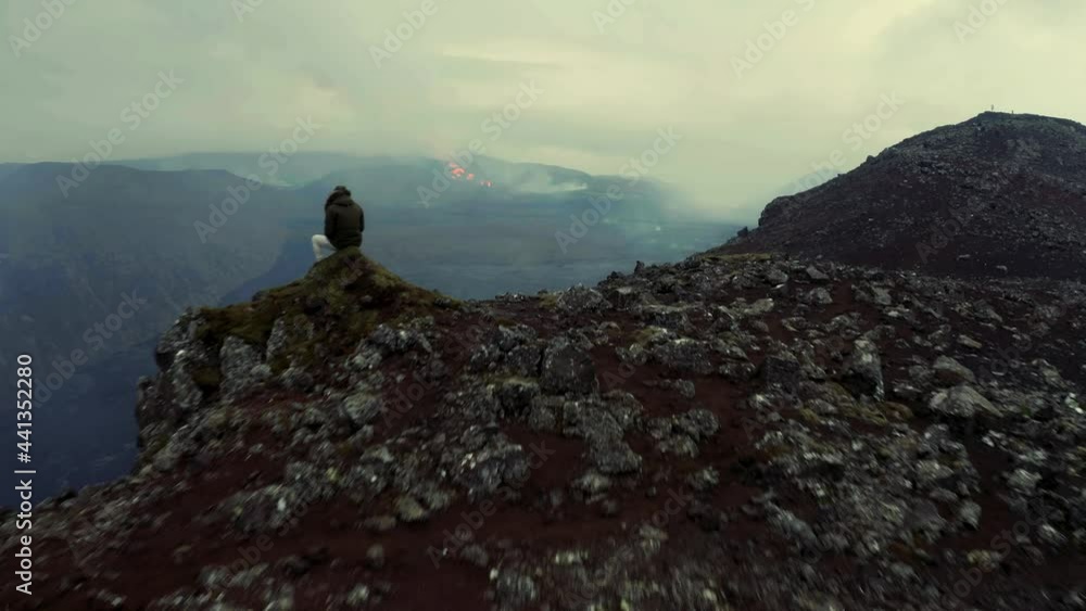 Aerial view over a person sitting on top of a mountain, looking down on a erupting Volcano, in Iceland - rising, drone shot Aerial view over a person sitting on top of a mountain, looking down on a erupting Volcano, in Iceland - rising, drone shot