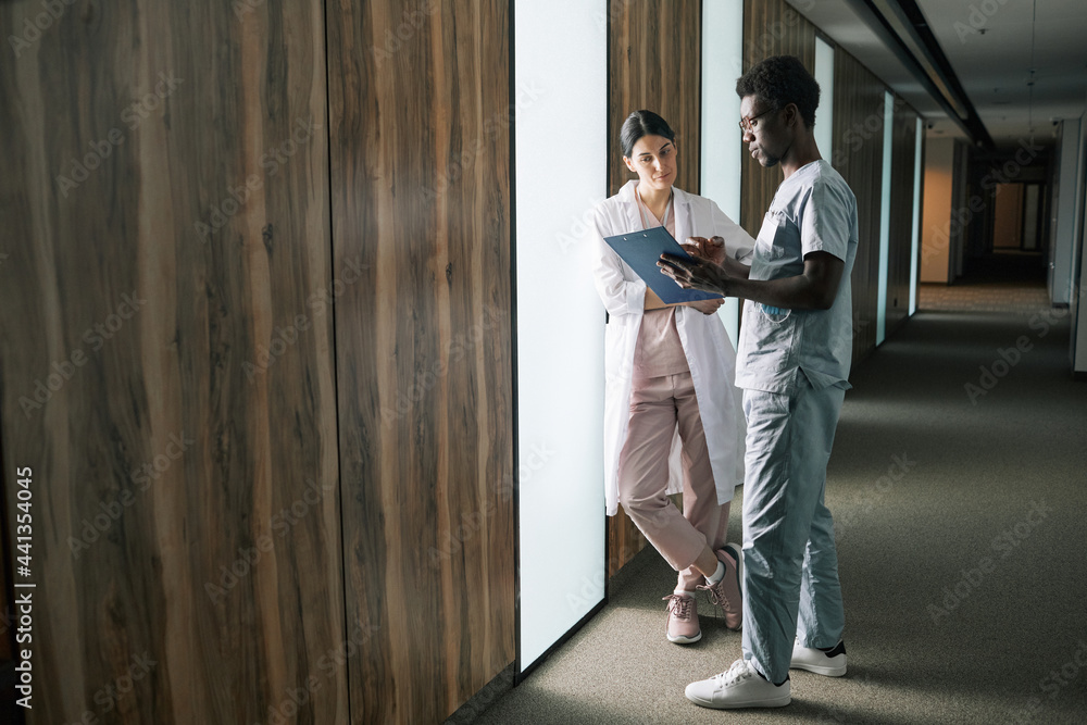 Full length portrait of young African-American medic talking to female colleague in clinic Full length portrait of young African-American medic talking to female colleague in clinic