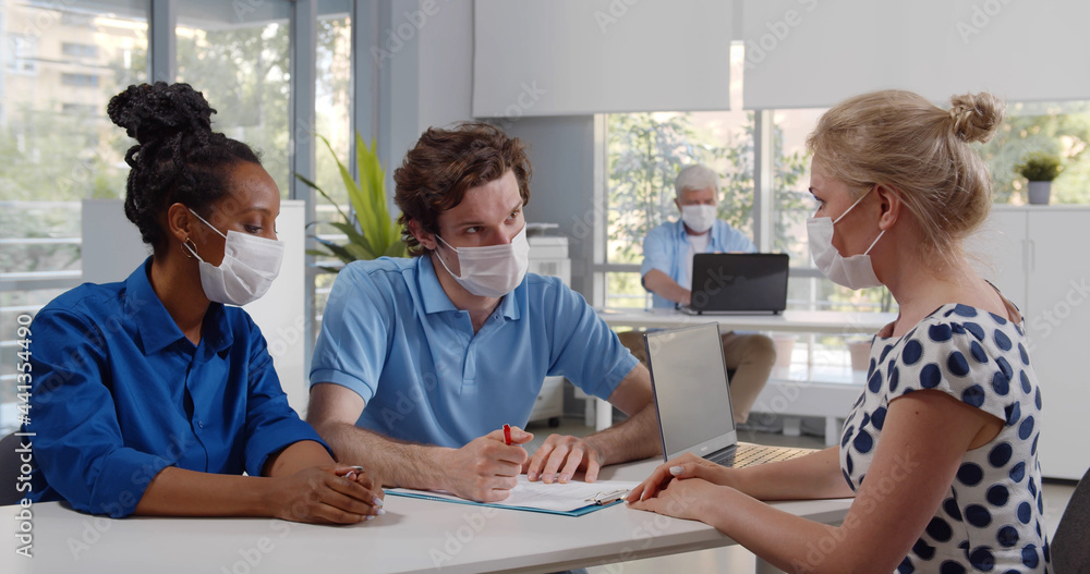 Fototapeta premium Young multiethnic couple in safety mask have meeting with realtor in office