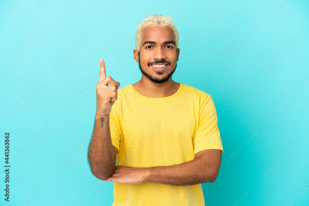 Young Colombian handsome man isolated on blue background happy and pointing up - obrazy, fototapety, plakaty 