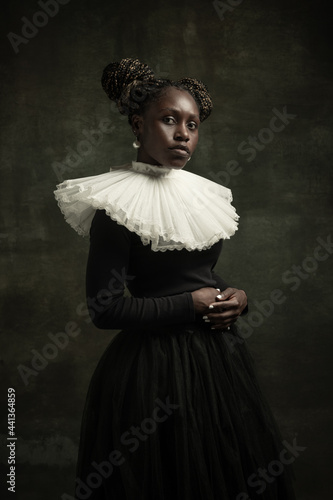 Fototapeta Portrait of medieval African young woman in black vintage dress with big white collar posing isolated on dark green background