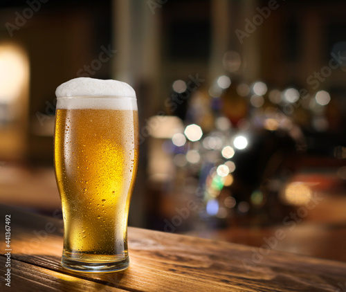 Cooled glass of pale beer with condensation drops on glass surface on the wooden table. Blurred bar at the background.
