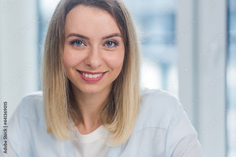 Close-up portrait a caucasian young blonde woman indoors looking at camera and smiling. Cute Happy Female in the office or at home with makeup and a beautiful smile and blue eyes. Closeup