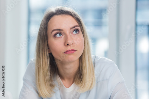 Close-up portrait of a young adult blonde business woman looking at the camera indoors at office or home on a blue window background. Face Confident female Manager in Formal Wear indoors