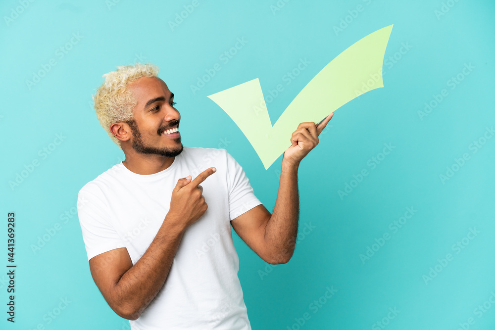 Young Colombian handsome man isolated on blue background holding a check icon and pointing it Young Colombian handsome man isolated on blue background holding a check icon and pointing it