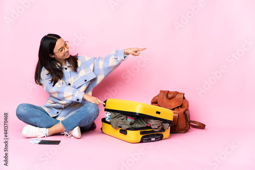 Traveler woman with a suitcase sitting on the floor pointing finger to the side and presenting a product