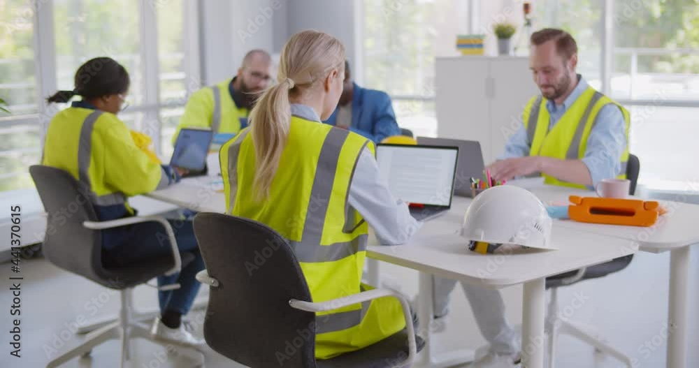 Young woman constructor wearing reflective vest posing at camera sitting at desk in modern office Young woman constructor wearing reflective vest posing at camera sitting at desk in modern office