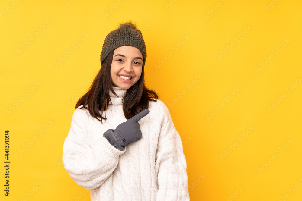 Young woman with winter hat over isolated yellow background pointing to the side to present a product Young woman with winter hat over isolated yellow background pointing to the side to present a product