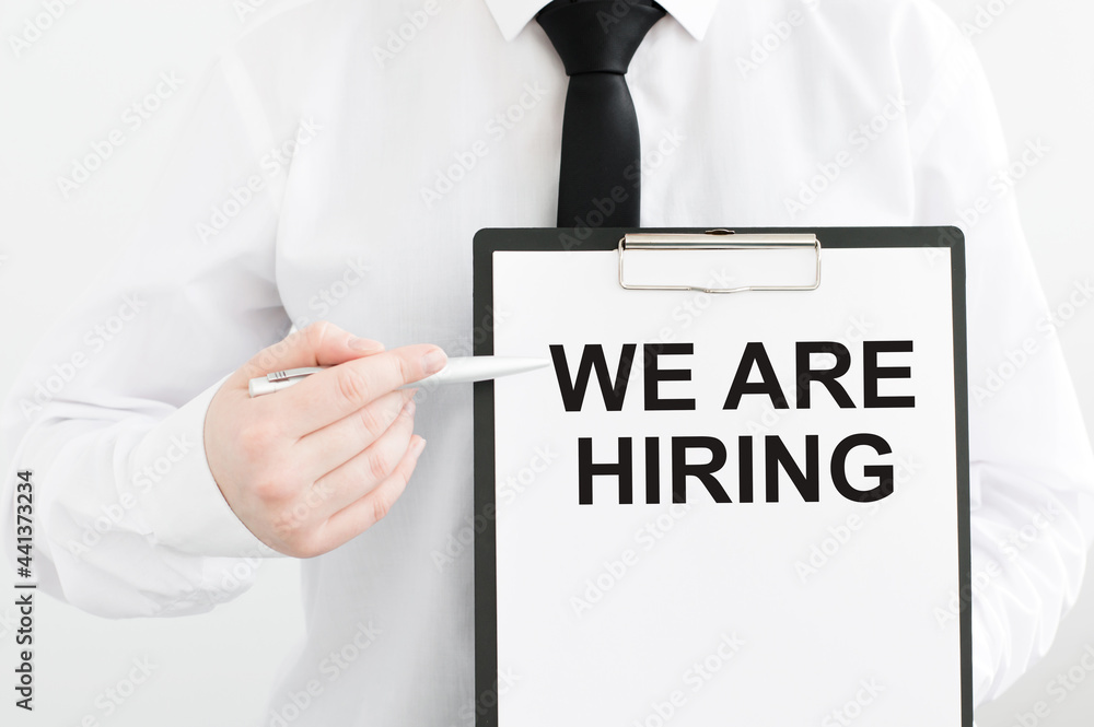 businessman holding a card with text WE ARE HIRING on a light background, points to the inscription with a pen businessman holding a card with text WE ARE HIRING on a light background, points to the inscription with a pen