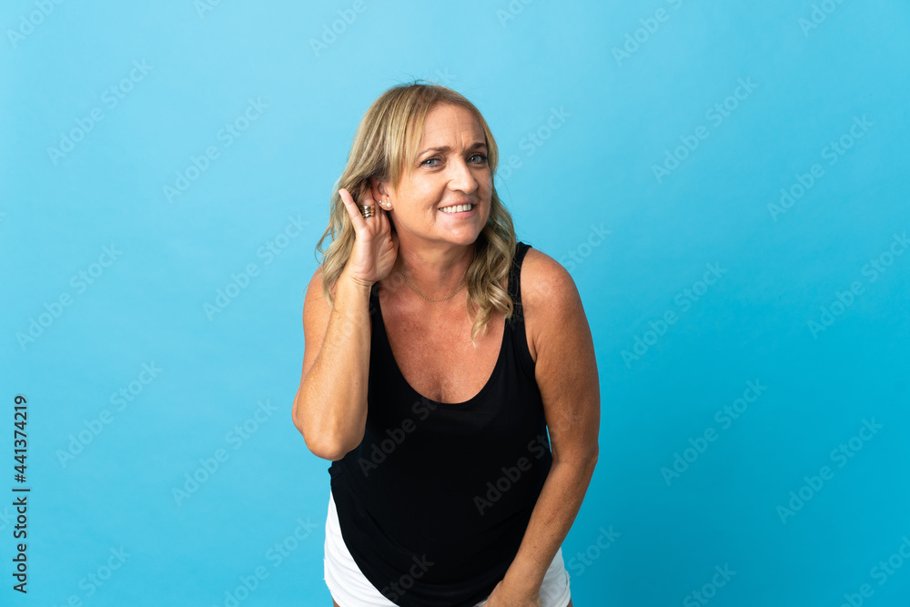 Middle aged blonde woman over isolated background listening to something by putting hand on the ear