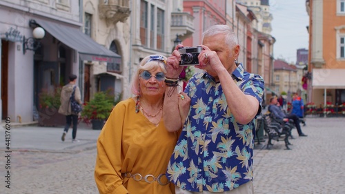 Elderly stylish couple tourists man and woman taking photos with old film camera, walking along city street. Senior travelers grandmother, grandfather enjoying time together. Summer vacation trip