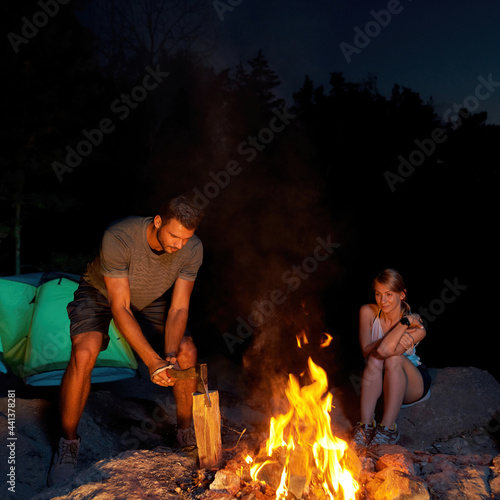 A couple of young tourists are camping in forest. Man chopping wood for a fire with an axe