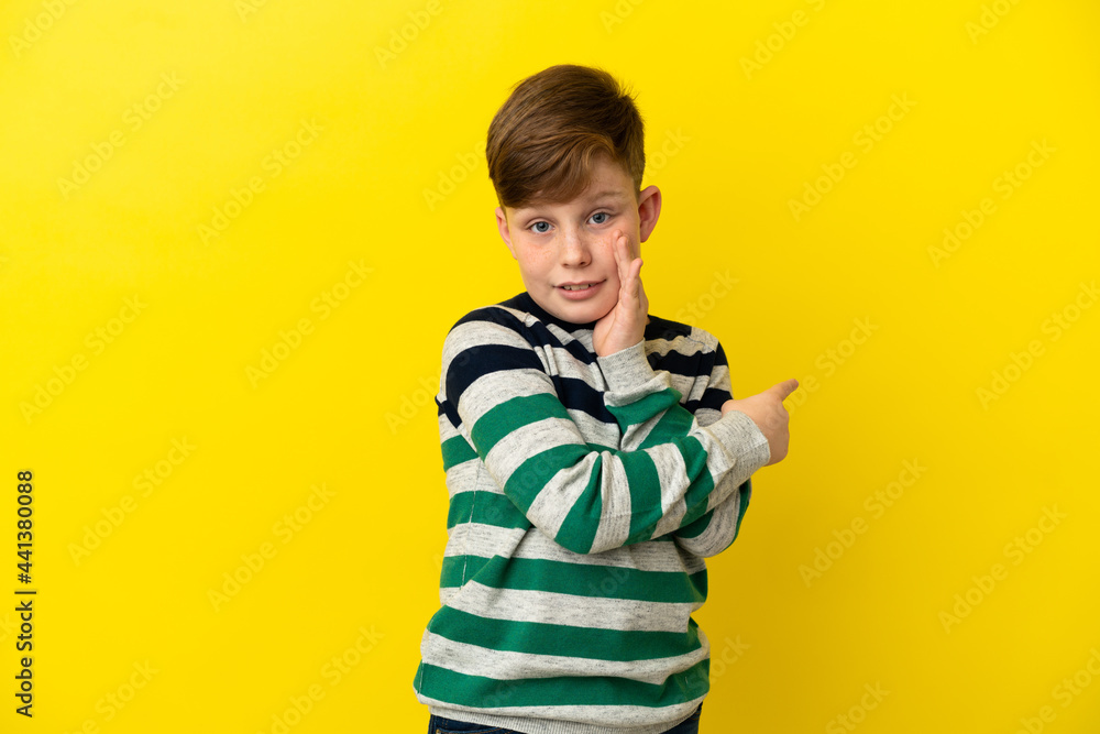 Little redhead boy isolated on yellow background pointing to the side to present a product and whispering something Little redhead boy isolated on yellow background pointing to the side to present a product and whispering something