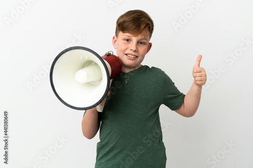 Little redhead boy isolated on white background holding a megaphone with thumb up