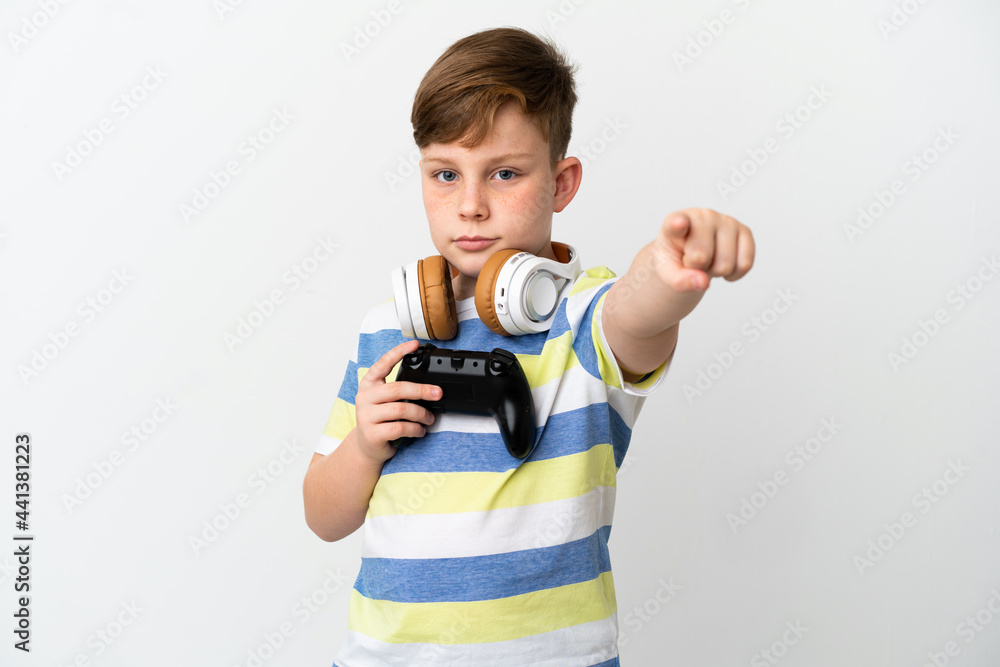 Little redhead boy holding a game pad isolated on white background surprised and pointing front Little redhead boy holding a game pad isolated on white background surprised and pointing front