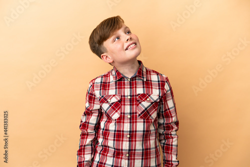 Little redhead boy isolated on beige background looking up while smiling