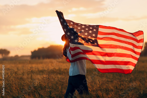 Woman farmer in wheat field waving american flag on sunset.