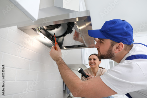 Worker repairing modern cooker hood and woman holding flashlight in kitchen
