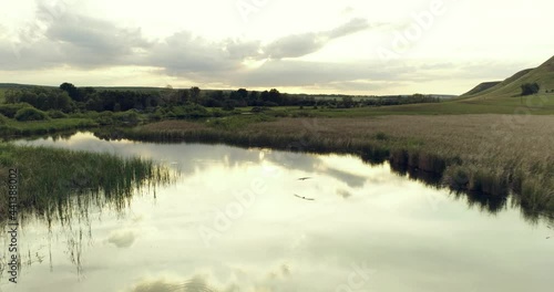 Stork flight on the river with beauty calm swampy backwater with reeds and thickets and bald hills background. Aerial above drone wide view at summer sunset.