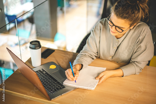 A woman in glasses studying online with her laptop, writing in notepad. Easy online education with computer and eLearning webinars concept