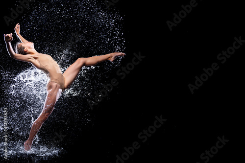 Wet young woman in tight beige swimsuit is dancing on floor under rain and splashes, drops of water. Girl modern dancer is doing tricks. Contemporary art dance. Freedom and freshness concept.