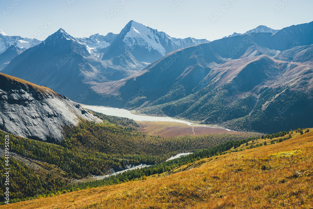 Obraz premium Wonderful alpine landscape with mountain lake and mountain river in valley with forest in autumn colors on background of snowy mountains silhouettes under blue sky. Beautiful mountain valley in autumn