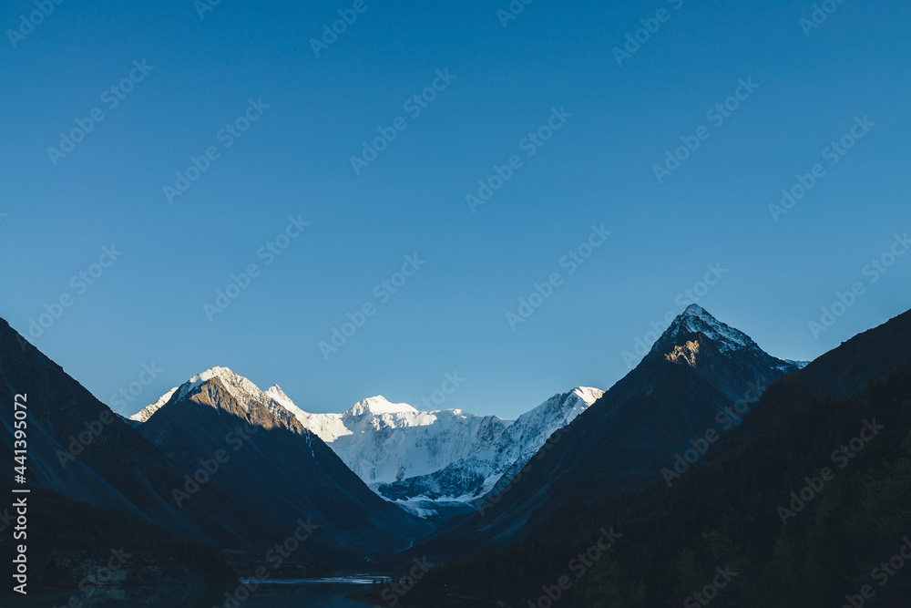 Awesome alpine landscape with mountains silhouettes and snow-covered mountain tops in gold sunshine under clear blue sky. Beautiful sunlit snow-white mountain wall and lake in shadowed highland valley Awesome alpine landscape with mountains silhouettes and snow-covered mountain tops in gold sunshine under clear blue sky. Beautiful sunlit snow-white mountain wall and lake in shadowed highland valley