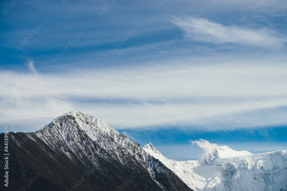 Atmospheric alpine landscape with snow-covered mountain top under cirrus clouds in blue sky. Awesome mountain scenery with beautiful pointy peak with snow and high snowy mountain wall with low clouds.