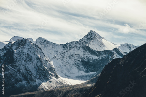 Great view to high snowy mountain wall with peaked top under cirrus clouds in sky. Alpine landscape with big snow covered mountains with sharp pinnacle in sunshine. White-snow pointy peak in sunlight.