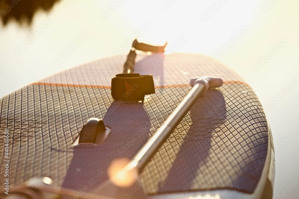 Fototapeta premium Paddle board and surf board with paddle in sunset light on water background close up. Surfing and SUP boarding equipment in sunset lights close-up. Outdoor water sports. Surfing lifestyle backgrounds.