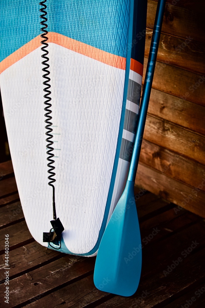Obraz premium Paddleboard and surfboard with paddle on brown wooden wall background close up. Surfing and SUP boarding equipment in sunset lights close-up. Outdoor water sports. Surfing lifestyle backgrounds.