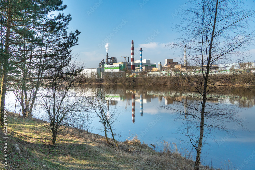 A large factory is located on the river bank. Toxic white smoke escapes from the factory's chimneys. The risk of an environmental disaster. Reflection in the water of the river of the plant