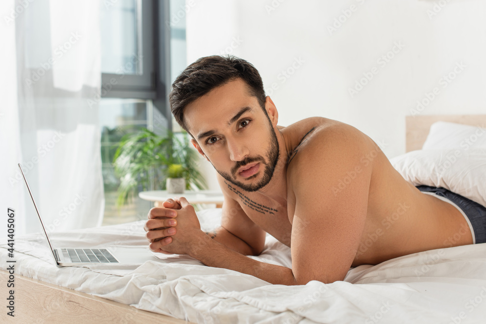 Shirtless man looking at camera near laptop on bed at home Shirtless man looking at camera near laptop on bed at home