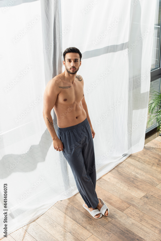 Obraz premium Shirtless man in pants and slippers looking at camera near window