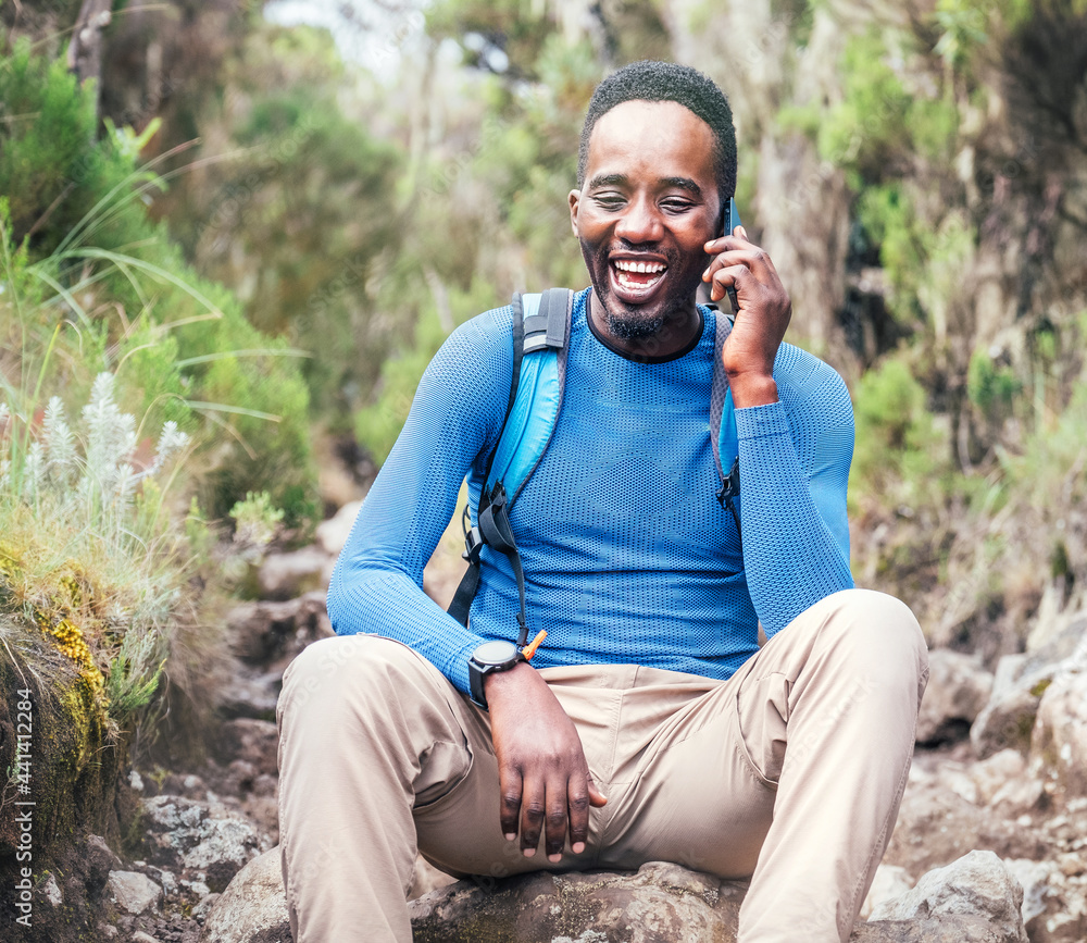 Φωτογραφία Young African ethnicity man talking via cellphone with somebody and cheerfully laughing as he having a hiking walk in the tropical forest