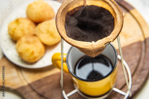 Café sendo coado no coador com uma caneca amarela abaixo com um prato de pão de queijo ao fundo. photo