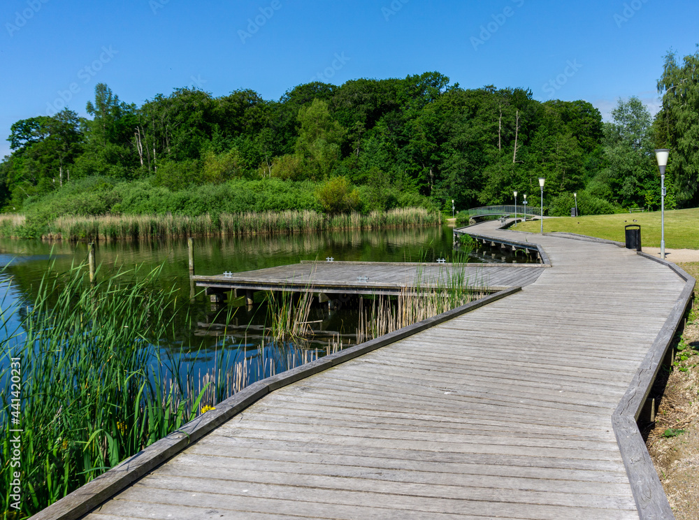 wooden boardwalk leads along the shores of a calm blue lake with a small bridge in the background